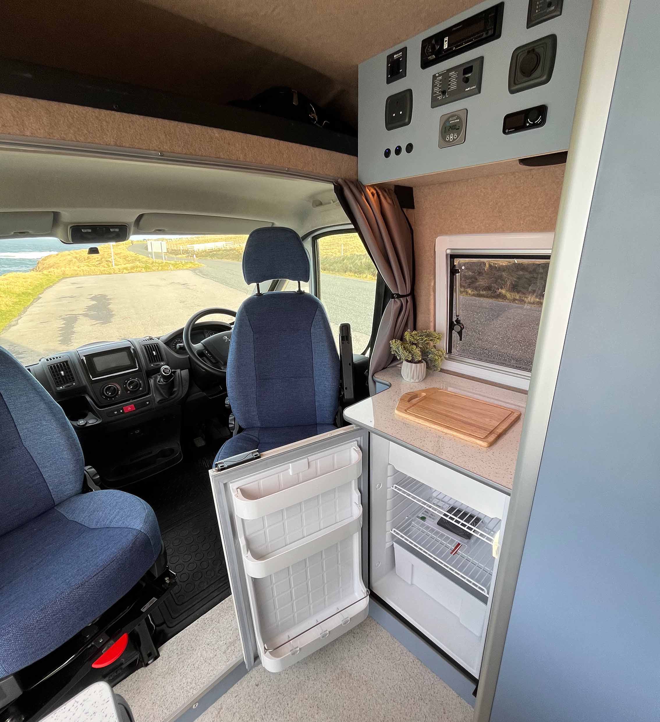 interior of van with the front seats swivelled towards the back, the fridge door open and the control panel up above